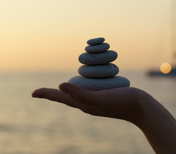 Hand holding a small stack of smooth dark stones against a soft sunset sky